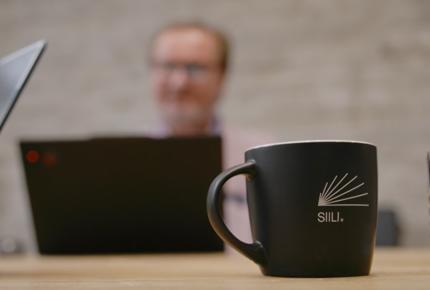Siili Solutions kuvituskuva. Siilin työntekijö kokoushuoneessa edessään Siilin logolla varustettu kahvikuppi. 