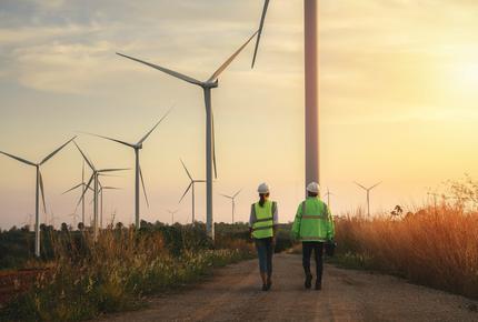 Back view of young maintenance engineers team working in wind turbine farm at sunset