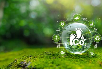 Sammalpinnalla oleva lasinen pallo, jossa näkyy hiilidioksidipäästöihin viittaava CO₂-symboli ja jalanjälkikuvio. Pallon ympärillä on kestävää kehitystä, kierrätystä, uusiutuvaa energiaa ja ympäristövastuuta kuvaavia ikoneita.